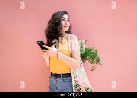 Young woman looking back while holding smart phone and standing with reusable mesh bag against pink wall Stock Photo