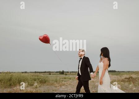 Bride and groom with heart shape balloon holding hands while walking against clear sky Stock Photo