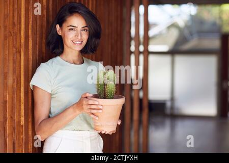Smiling female entrepreneur holding cactus plant while standing by wooden wall in office Stock Photo