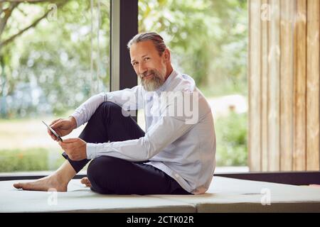 Smiling bearded man using digital tablet while relaxing on bed by window at home Stock Photo