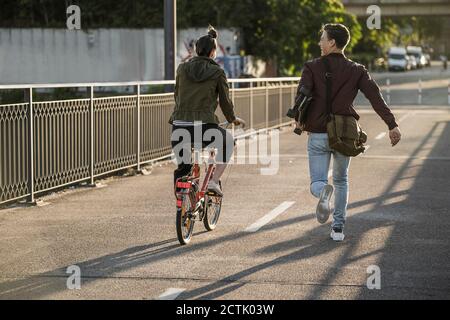 Young man carrying skateboard while running by girlfriend riding bicycle in city Stock Photo