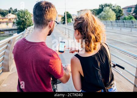Couple using smart phone while standing with bicycles on bridge at sunset Stock Photo