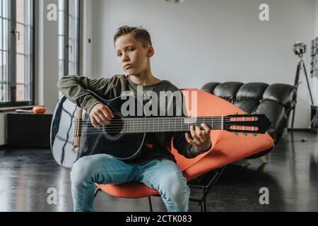 Boy playing guitar while sitting on chair at home Stock Photo