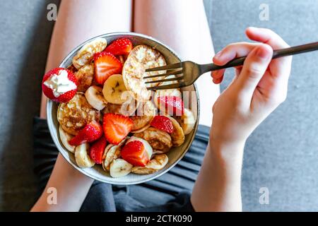 Hands of teenage girl eating bowl of mini pancakes with strawberries and bananas Stock Photo