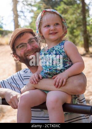 Father holding daughter in arm while sitting on rod in public park Stock Photo