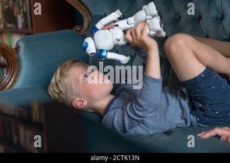 Cute boy playing with robot while lying on sofa in living room at home Stock Photo