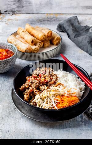 Bowl of fried sesame beef, salad and rice with dipping sauce and spring rolls in background Stock Photo