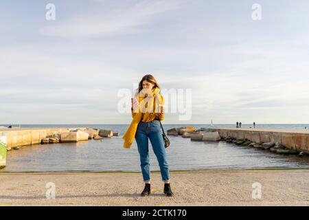 Thoughtful woman using mobile phone while holding coffee cup against sea Stock Photo