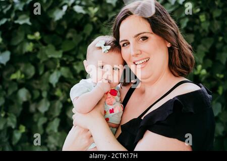 Smiling mother holding baby girl against ivy Stock Photo