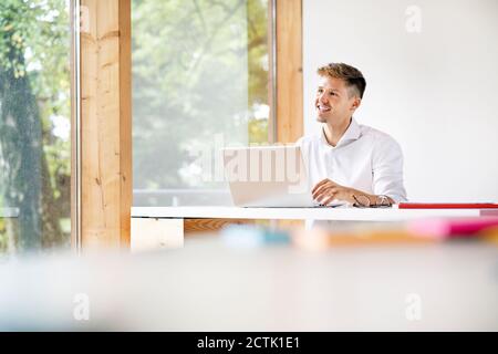 Smiling businessman with laptop on desk contemplating while sitting in office Stock Photo