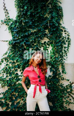 Woman with hands in pockets standing against wall covering with ivy at Aflama, Lisbon, Portugal Stock Photo