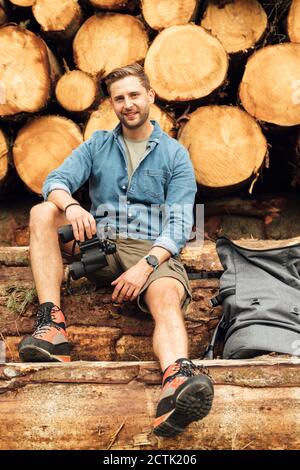 Smiling man with binoculars and backpack sitting on log against woodpile Stock Photo