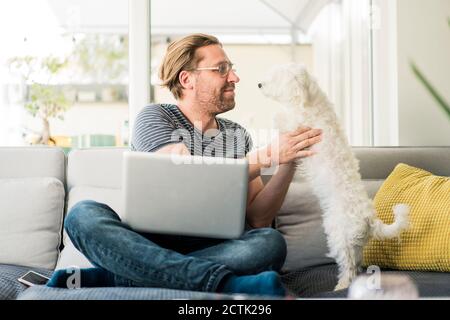 Man with laptop holding dog while sitting on sofa Stock Photo