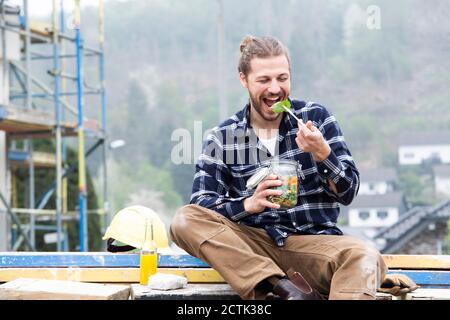 Construction worker eating salad while sitting outdoors at construction site Stock Photo