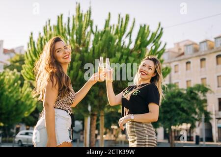 Cheerful female friends toasting beer bottles while standing in city Stock Photo