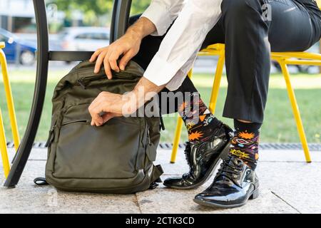 Male entrepreneur wearing colorful socks zipping backpack while sitting on chair at sidewalk cafe Stock Photo