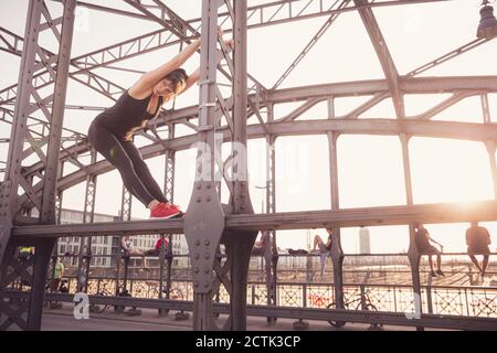 Woman stretching while exercising on metal bridge in city at sunset Stock Photo