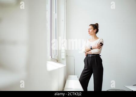 Thoughtful businesswoman with smart phone looking through window Stock Photo