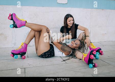 Cheerful female friends with roller skates relaxing on footpath against wall in city Stock Photo
