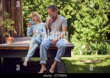 Father and daughter brushing teeth while sitting in yard Stock Photo