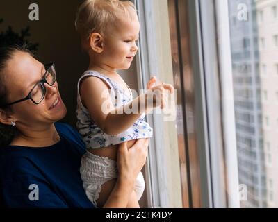 Smiling mother with baby boy looking through window at home Stock Photo