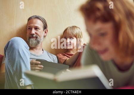 Woman reading book while father and daughter looking away in background at home Stock Photo