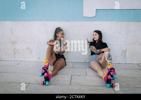 Friends with roller skates relaxing while sitting on footpath against wall in city Stock Photo