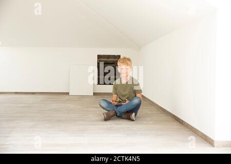 Smiling boy holding smart phone while sitting on floor in attic of new home Stock Photo