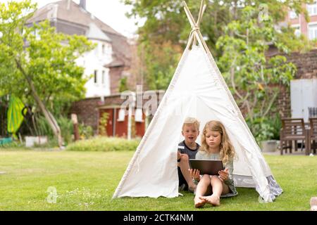 Cute siblings with digital tablet sitting in tent on grass at back yard during weekend Stock Photo
