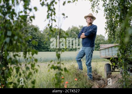 Thoughtful man wearing hat with arms crossed standing in yard Stock Photo