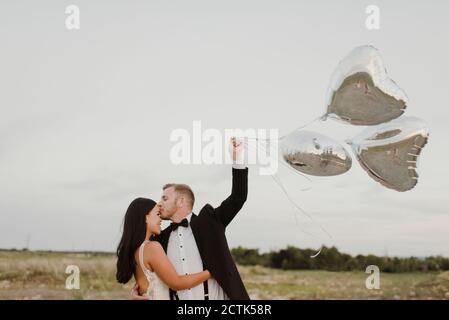 Smiling groom with heart shape balloons kissing bride on forehead against sky Stock Photo