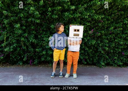 Boy looking at friend wearing robot mask while standing against plants on road in park Stock Photo