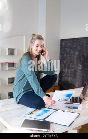 Smiling woman sitting on desk in home office talking on the phone Stock Photo