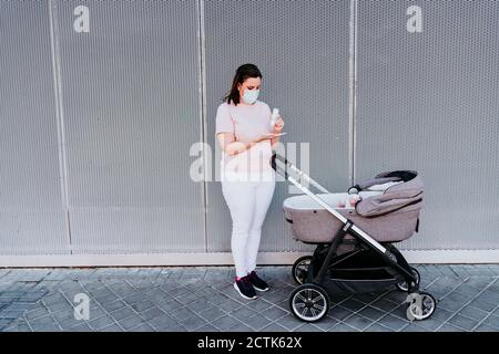 Mother wearing protective mask, using hand sanitizer, baby girl in stroller Stock Photo