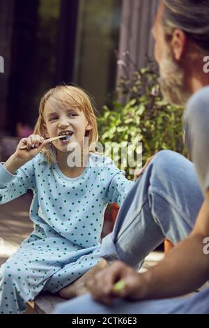 Girl looking at father while brushing teeth with him in yard Stock Photo