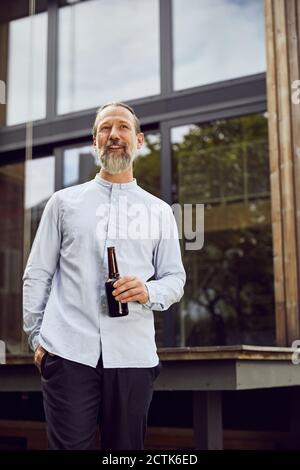 Bearded mature man holding beer bottle while standing against tiny house Stock Photo