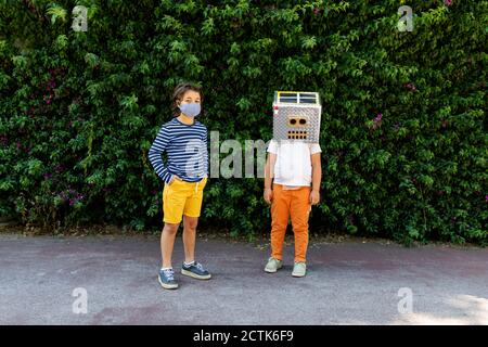 Boy in face mask standing with friend wearing robot costume against plants in park Stock Photo