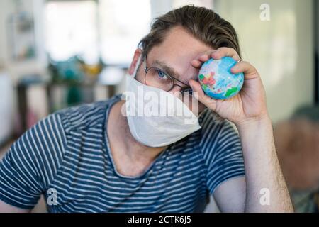 Worried man with globe wearing face mask while sitting at home office Stock Photo