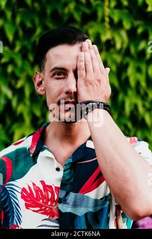 Man covering face with one hand while standing against ivy hedge Stock Photo