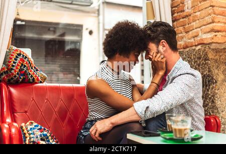 Cheerful woman pulling boyfriend's cheeks while sitting on sofa in cafe Stock Photo