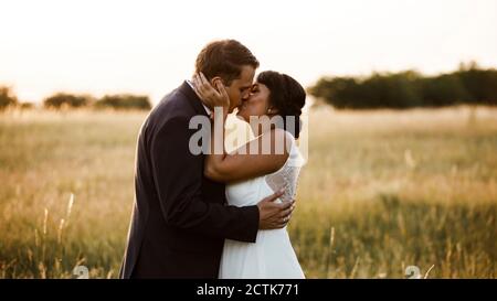 Affectionate young bridal couple kissing at field during sunset Stock Photo