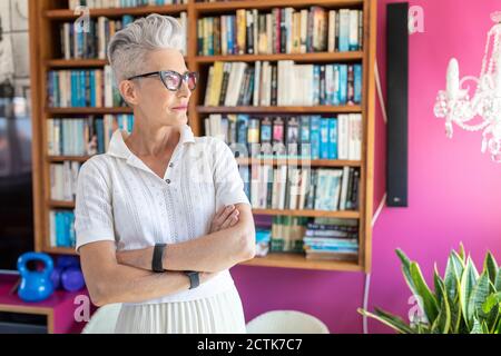 Senior female with arms crossed standing against bookshelf at home Stock Photo