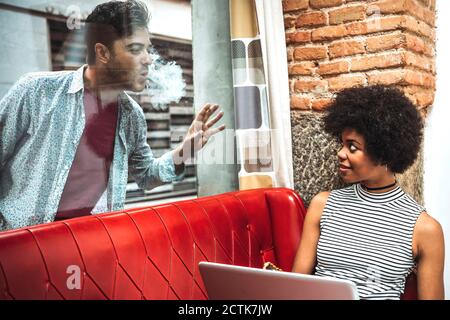Man emitting smoke from mouth while looking at girlfriend sitting in cafe through window Stock Photo