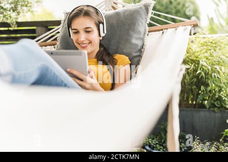 Smiling young woman wearing headphones using digital tablet while relaxing on hammock in yard Stock Photo