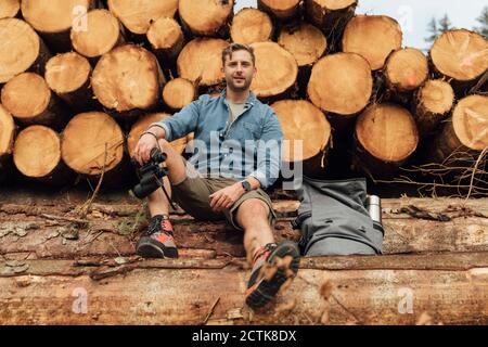 Man with binoculars and backpack sitting on log against woodpile in forest Stock Photo