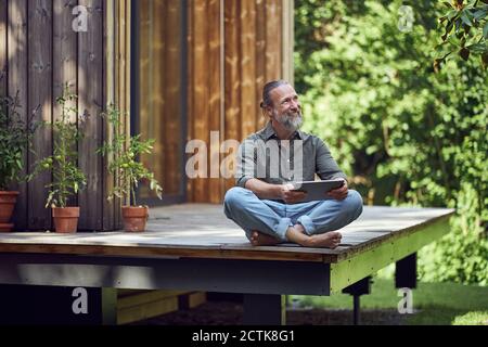 Bearded mature man holding digital tablet contemplating while sitting outside tiny house Stock Photo