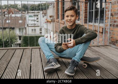 Smiling boy using smart phone while sitting on skateboard against railing Stock Photo