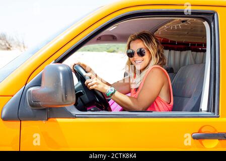 Smiling blond woman sitting in van and hands on steering wheel Stock Photo