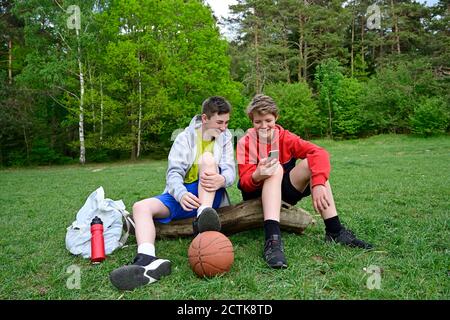 Smiling boys using mobile phone while sitting on log in forest Stock Photo