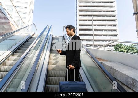 Businessman with suitcase using smart phone while standing on escalator in city Stock Photo
